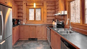 A log cabin kitchen with wooden cabinets, a stainless steel refrigerator, a dishwasher, a sink, and a window overlooking a snowy landscape.