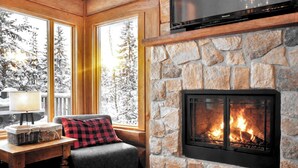Ein gemütliches Zimmer mit Kamin aus Stein, einer Couch mit rotem und schwarzem Karokissen, einem Fernseher, der über dem Kamin angebracht ist, und einem Fenster mit Blick auf schneebedeckte Bäume.