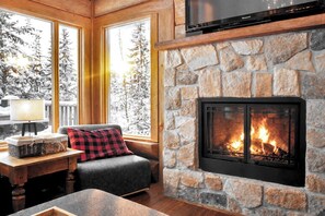 A cozy room with a stone fireplace, a sofa with a red and black checkered pillow, a television mounted above the fireplace, and a window with a view of snow-covered trees.