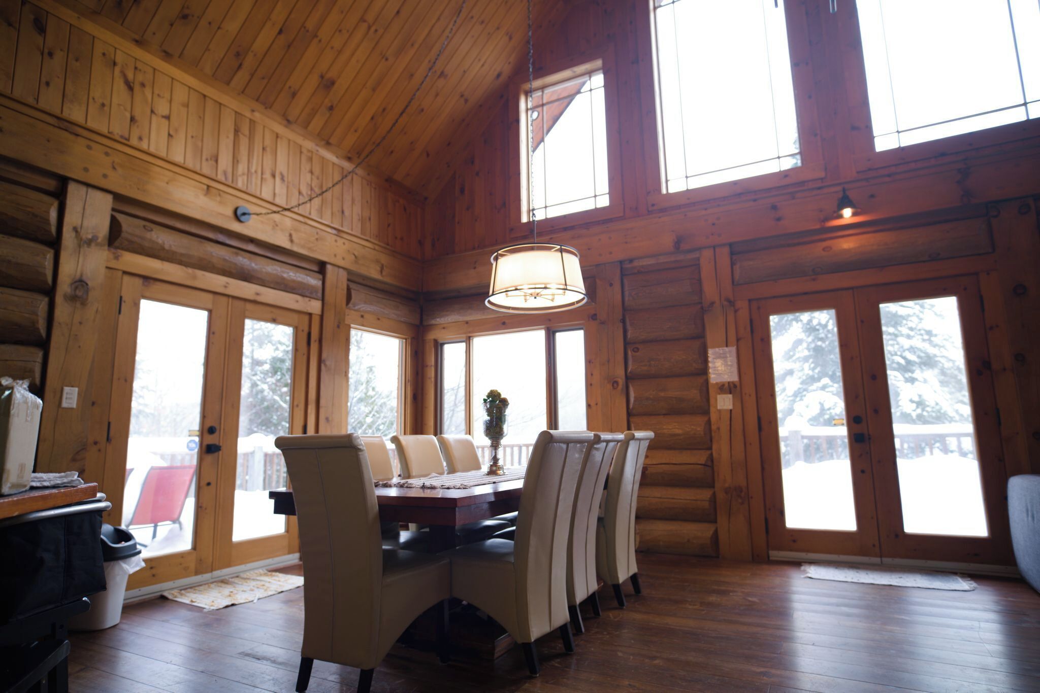 A log cabin interior with a dining table, chairs, a chandelier, and large windows.