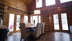 Ein Holzblockhaus-Interieur mit Esstisch, Stühlen, einer Chandelier und großen Fenstern.