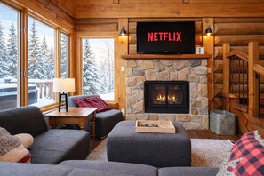 A cozy room with a fireplace, a television mounted above it, and a view of snow-covered trees outside. - Belvedere du Lac Lodge (Sainte-Adèle)