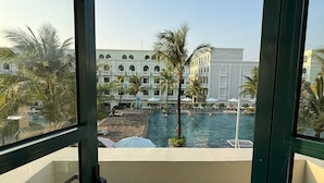 Blick von einem Balkon auf einen Swimmingpool, Palmen und ein großes Gebäude mit mehreren Balkonen.