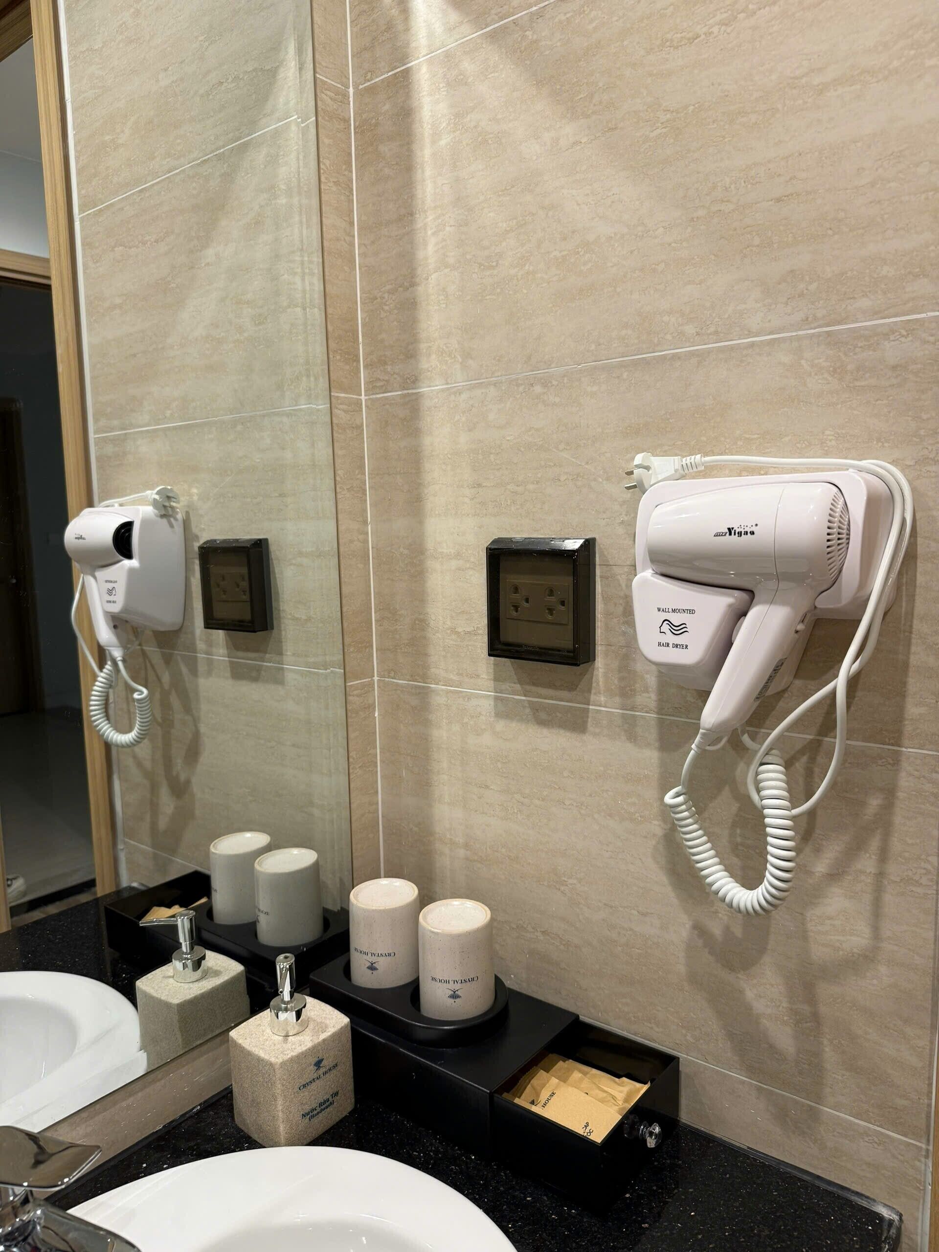 A bathroom with a mirror, two hair dryers mounted on the wall, a sink, and toiletries.