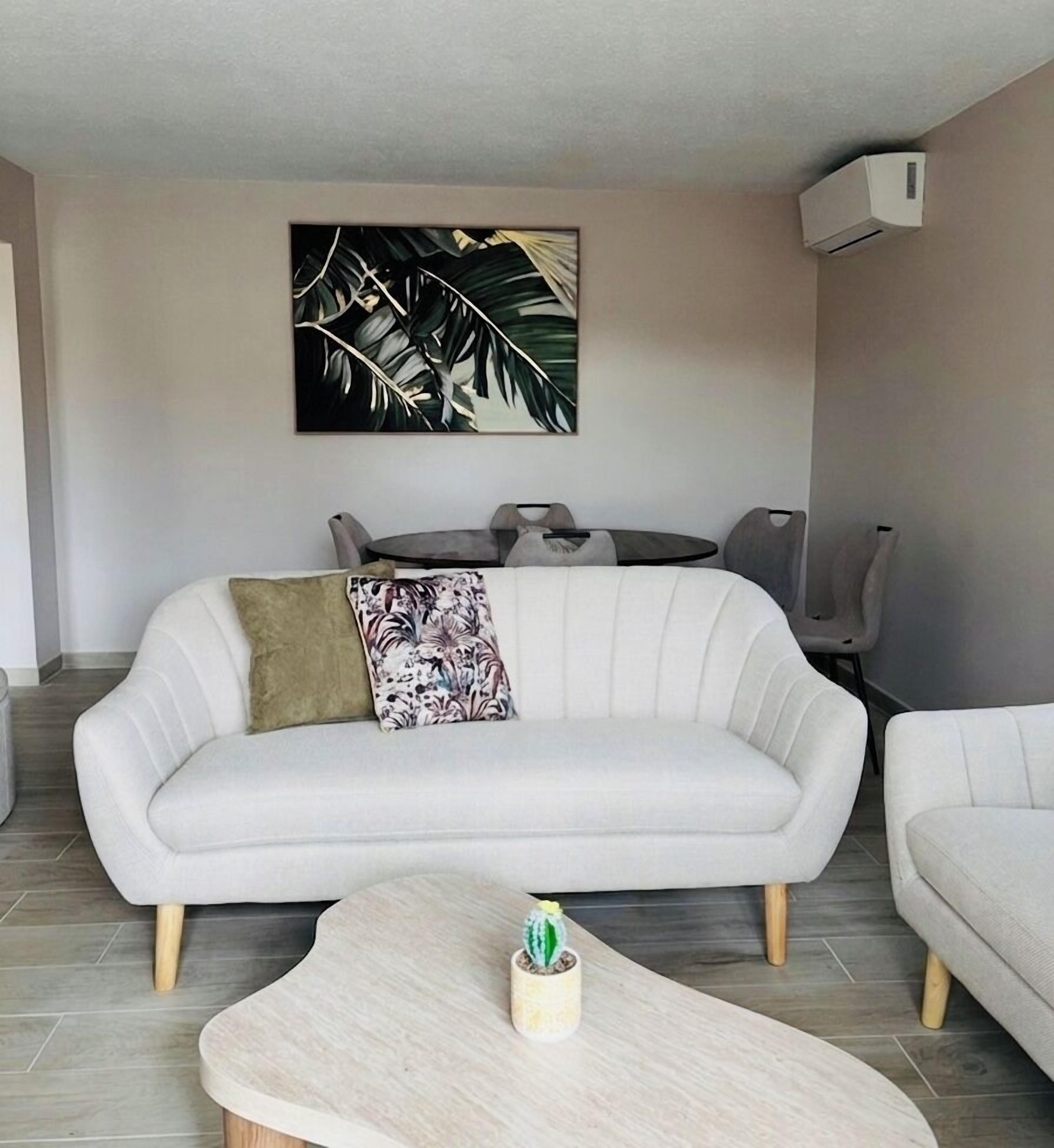 A modern living room with a white sofa, a coffee table, a dining table with chairs, and a large plant on the wall.