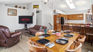 Een woonkamer met een televisie, een bruine leren bank, een houten eettafel voor vier personen, en een keuken met houten keukenkastjes.