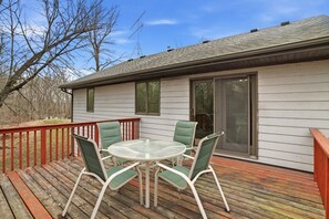 A deck with a glass table and chairs, a sliding glass door, and a white exterior wall.
