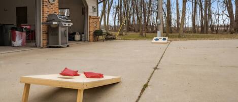 Eine Beton-Terrasse mit einem Holztisch, roten Kissen, einem Grill und einer Garage mit einer Uhr.