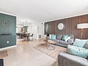 Ein modernes Wohnzimmer mit einem grauen Sofa, einem Couchtisch und einem Essbereich, im Hintergrund ist eine Küche erkennbar.
