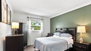 Quarto com cama, mesas de cabeceira, cômoda, janela com cortinas e uma obra de arte na parede.