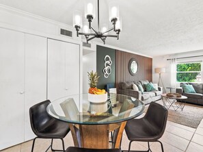 A modern living room with a glass dining table, black chairs, a grey sofa, and a wooden accent wall. - Baywood Colony - 2BR 2BA - Siesta Key Area (Sarasota)