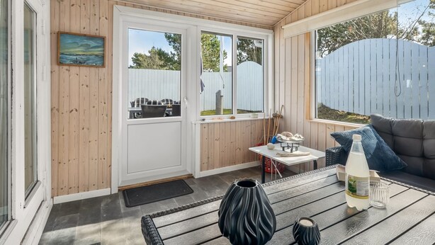 Eine überdachte Terrasse mit Holzboden, einer Couch, einem Tisch mit Flasche und Kerze sowie einem Gemälde an der Wand.