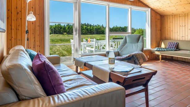 Ein gemütliches Zimmer mit Holzboden, einer Couch mit Kissen, einem Sessel, einem Tisch und einem großen Fenster mit Blick auf Grünflächen.