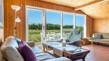 Et hyggeligt rum med træloft, en sofa med puder, en stol, et bord og et stort vindue med udsigt over grøn vegetation.