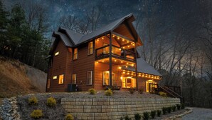 A two-story log cabin with a balcony, surrounded by a stone retaining wall and small shrubs. - New Pool Cabin! Luxury Design, Game Rm, Hot Tub! (Sevierville)