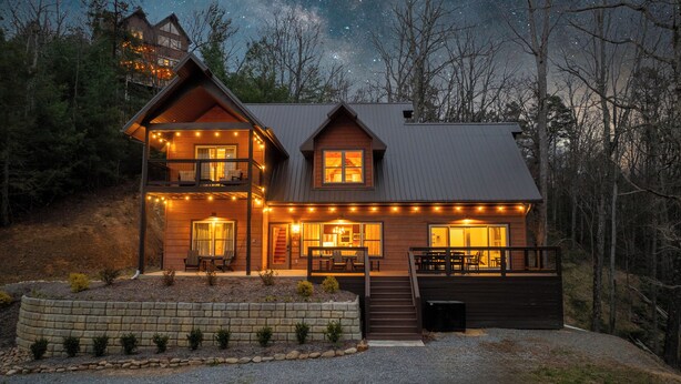 A two-story log cabin with a balcony, string lights, and a stone retaining wall. - New Pool Cabin! Luxury Design, Game Rm, Hot Tub! (Sevierville)