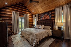 A log cabin bedroom with a large bed, wooden walls, a ceiling fan, and a painting above the bed. - New Pool Cabin! Luxury Design, Game Rm, Hot Tub! (Sevierville)