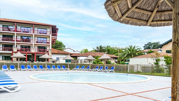 Un área de piscina con sillas de descanso, un edificio con balcones y una estructura de techo de paja.