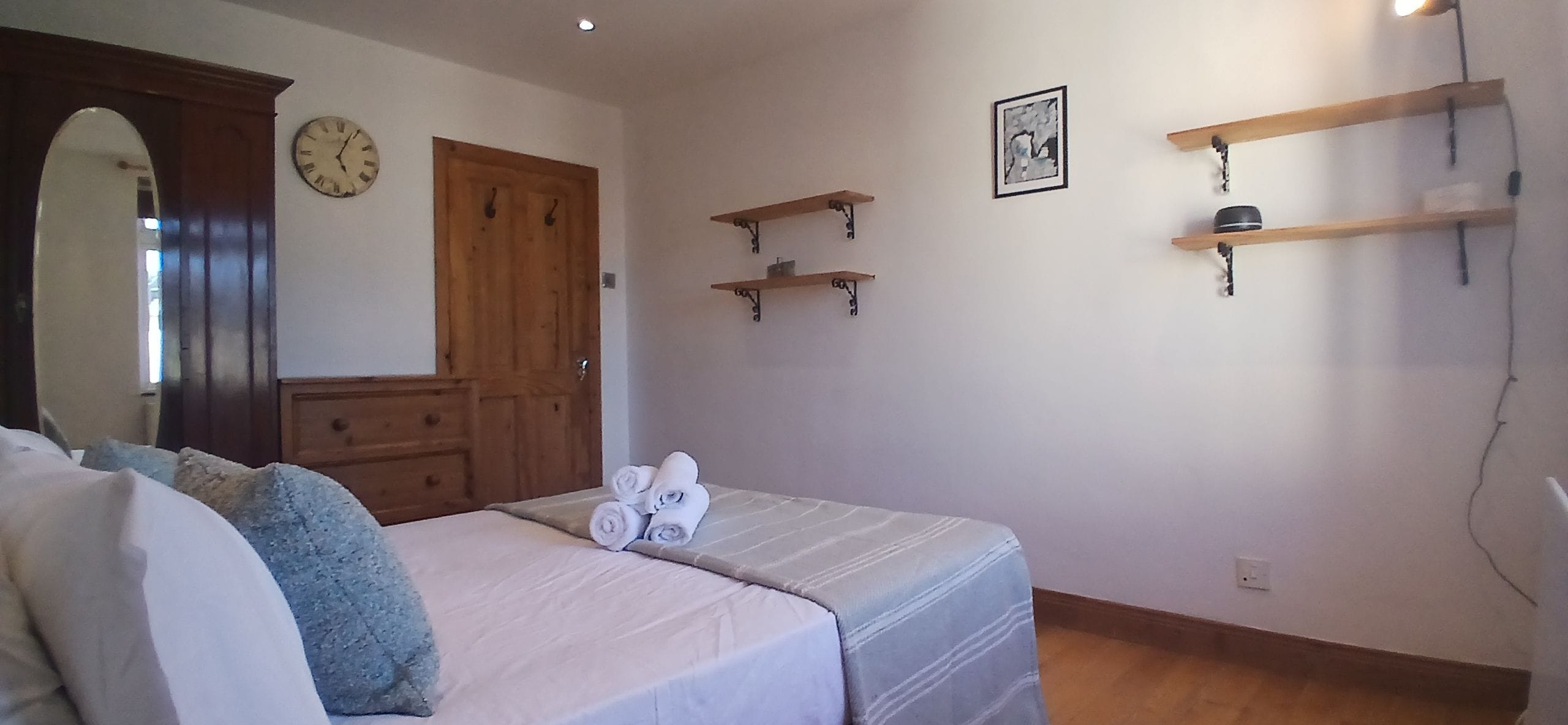 A bedroom with a bed, a dresser, a mirror, a clock, and two wall-mounted shelves.