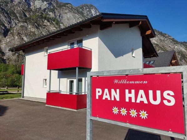 A building with a red and white facade, a sign reading 'Willkommen PAN HAUS,' and a mountainous backdrop. - Pan Haus Bad Goisern (Bad Goisern)