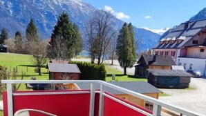 A balcony with a glass table and chairs, a red railing, and a view of mountains and houses.