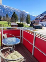 Un balcón con una mesa y sillas de vidrio, barandilla roja y vistas a montañas y casas.