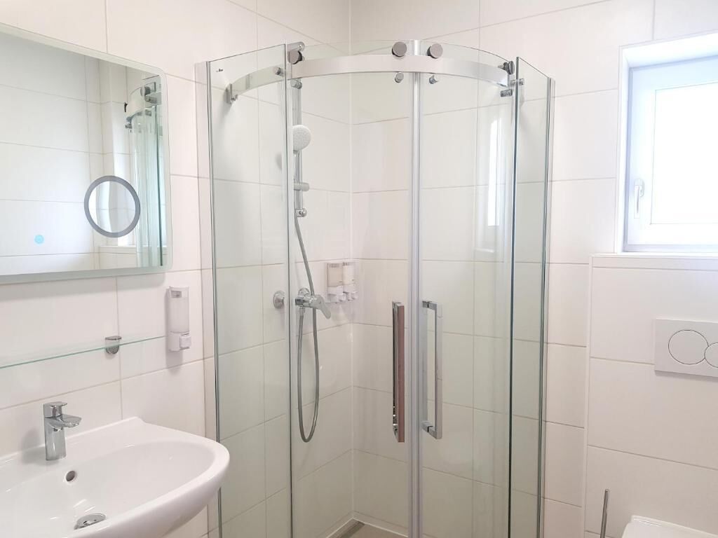 A glass-enclosed shower with a handheld showerhead, a white sink with a mirror above it, and a window with blinds.