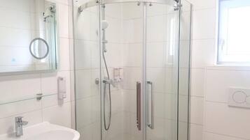 A glass-enclosed shower with a handheld showerhead, a white sink with a mirror above it, and a window with blinds.