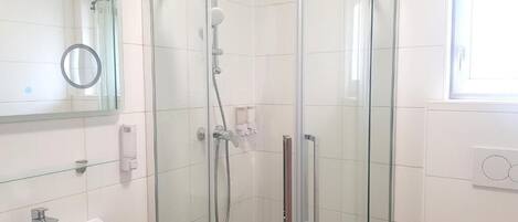 A glass-enclosed shower with a handheld showerhead, a white sink with a mirror above it, and a window with blinds.