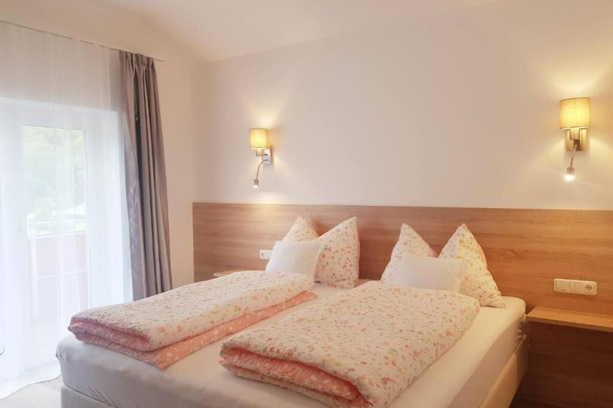 A double bed with a wooden headboard, two wall-mounted lamps, and a window with curtains.