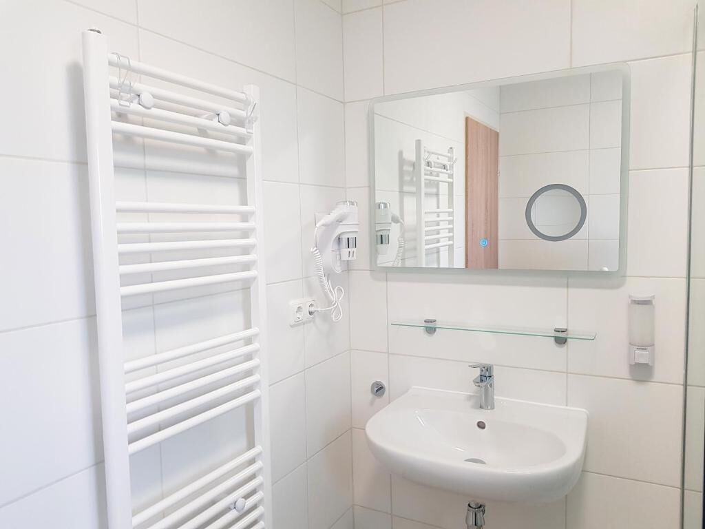 Un baño con un radiador blanco, un estante de vidrio sobre un lavabo y un espejo que refleja una puerta y un objeto circular.