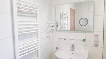 Une salle de bain avec un radiateur blanc, une étagère en verre au-dessus d’un lavabo et un miroir qui reflète une porte et un objet circulaire.