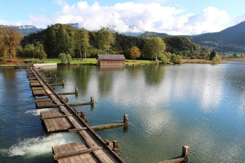 Eine Holzstege führt in einen ruhigen See mit Bergblick und einem kleinen Gebäude am Ufer.