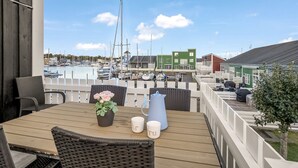 A wooden deck with outdoor seating, a view of a marina with boats, and a green building in the background. - "Björk" - 10m from the sea by Interhome (Ebeltoft)