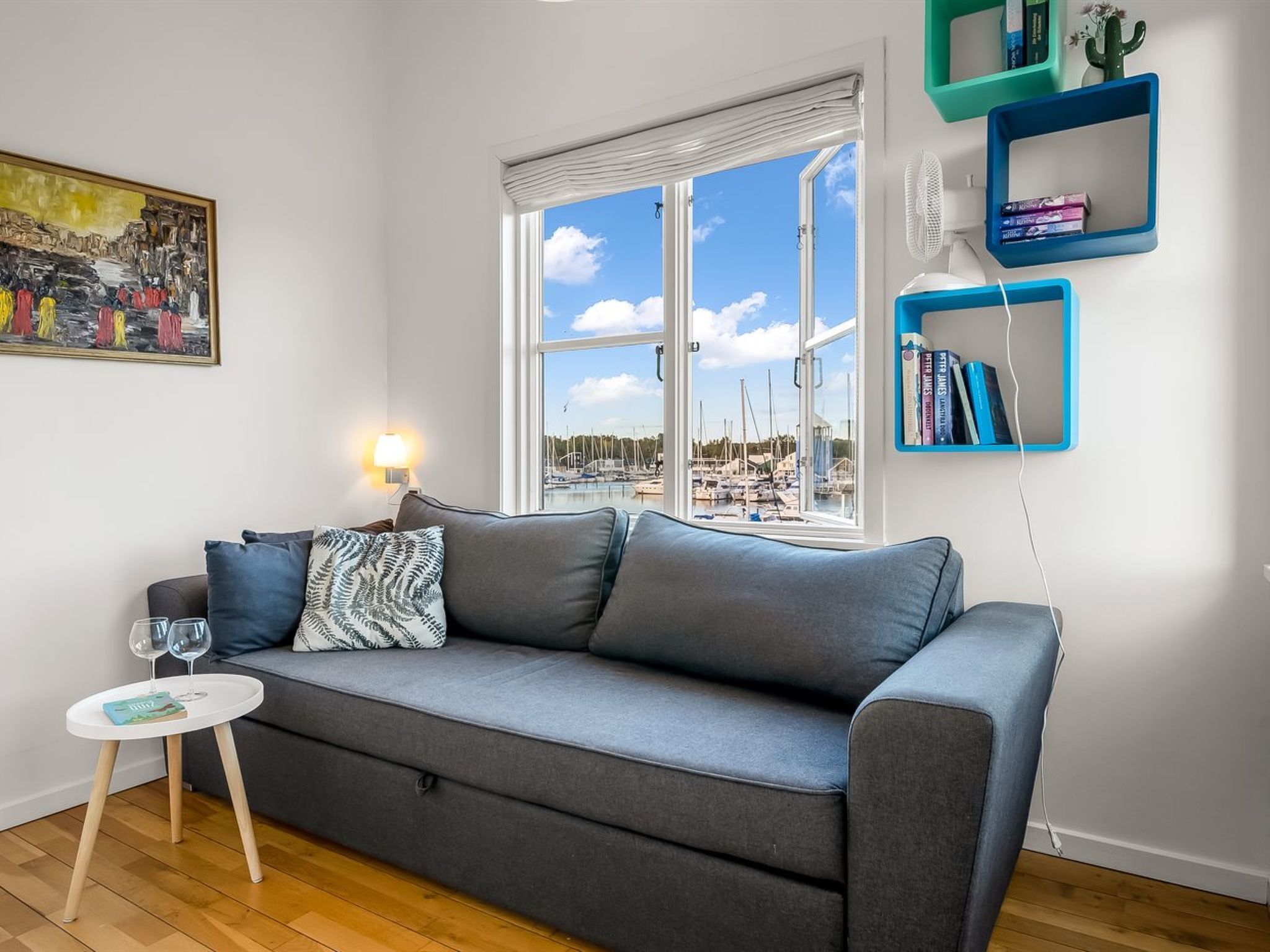 Een woonkamer met een grijze bank, een klein wit bijzettafeltje, een schilderij in een lijst en uitzicht op een jachthaven door een groot raam.