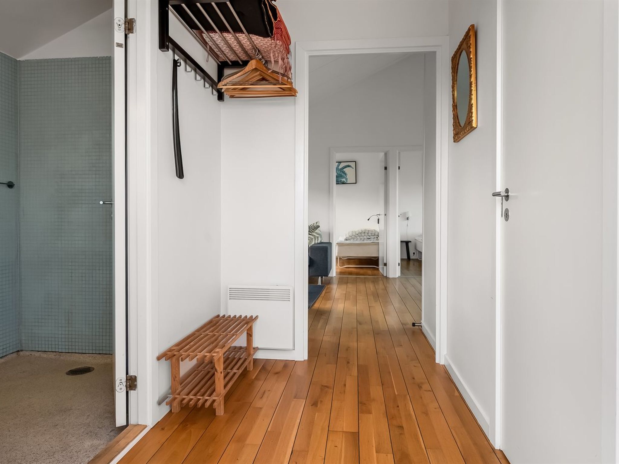 A hallway with wooden flooring, a bench, a drying rack, and a bed in the distance.