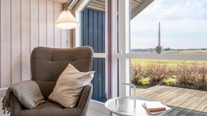 Ein gemütliches Zimmer mit einem großen Fenster, einem Sessel mit Kissen, einem kleinen Tisch mit Tasse und Buch sowie einer Hängelampe.