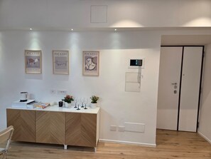 A modern room with a wooden herringbone cabinet, a small table with a kettle, wine glasses, and a potted plant. There are three framed artworks on the wall and a door with a handle. - Clodio 13 – Cozy Apartment in the Heart of Rome (Roma)