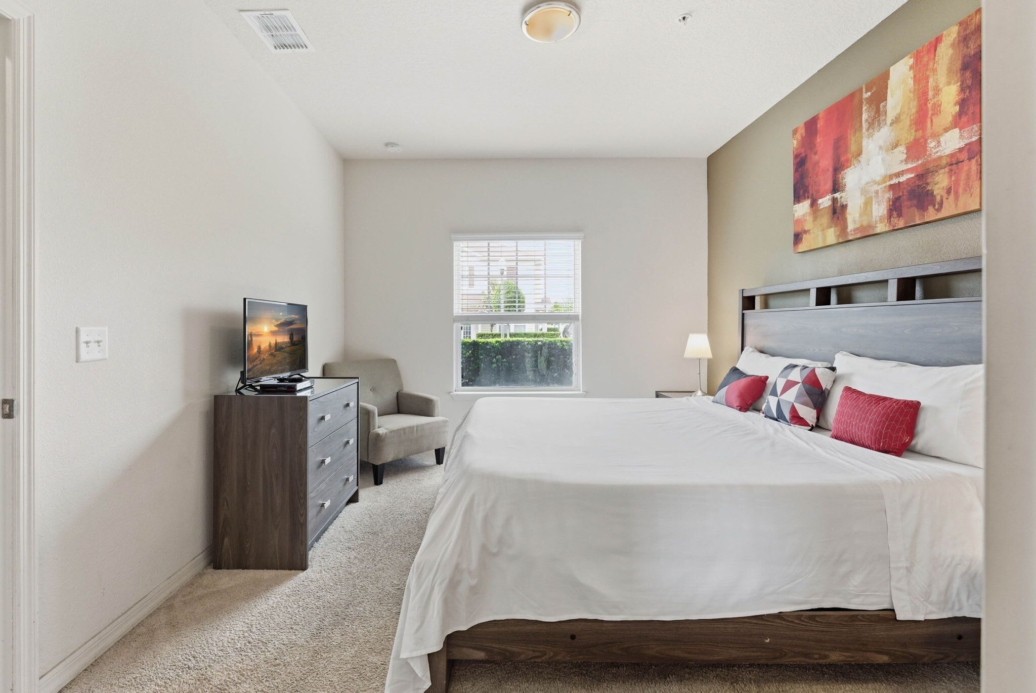 A bedroom with a bed, dresser, chair, window, and artwork.