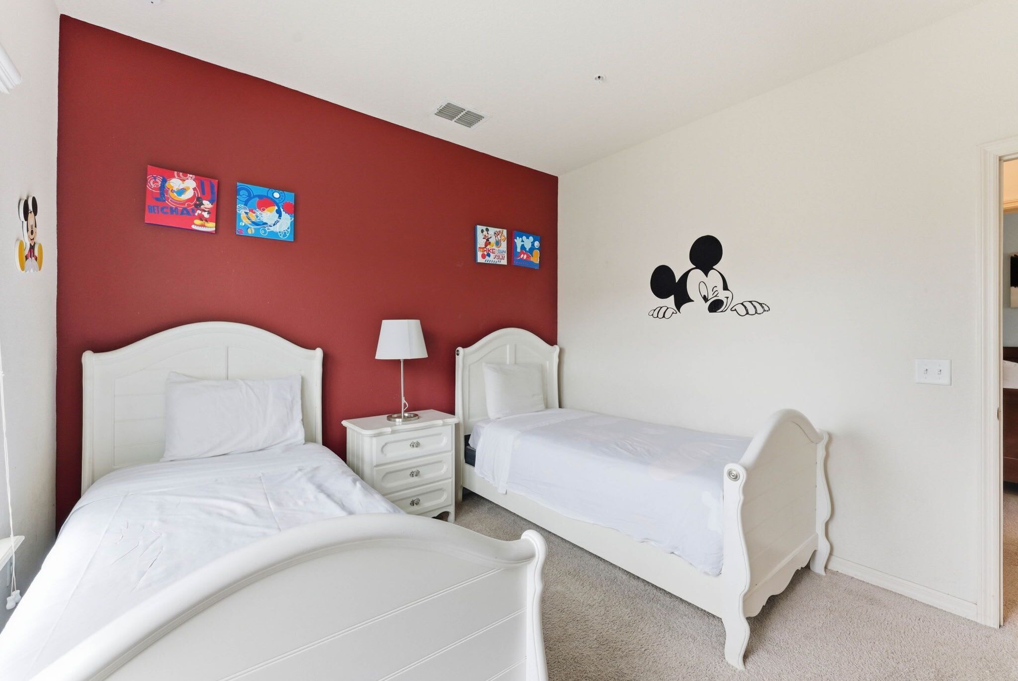 Un dormitorio con dos camas, una mesita de noche, una lámpara y detalles decorativos en la pared, incluyendo a Mickey Mouse y obras de arte coloridas.