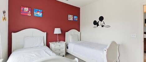 Un dormitorio con dos camas, una mesita de noche, una lámpara y detalles decorativos en la pared, incluyendo a Mickey Mouse y obras de arte coloridas.