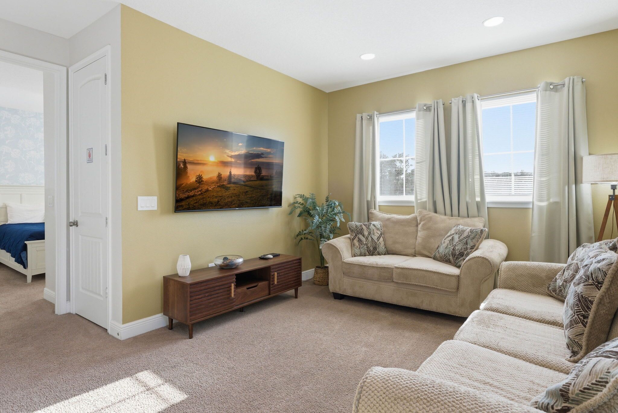 Una sala de estar con un sofá color beige, un televisor de pantalla plana montado en la pared, una ventana con cortinas y una puerta que da a otra habitación.