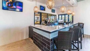 Eine Bar mit einer Theke, Barhocker, hängenden Leuchten und einem an der Wand befestigten Fernseher.