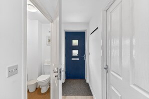 A narrow hallway leading to a room with a blue door, white walls, a white toilet, and a small rug. - Stylish House • Sleeps 5 • Driveway Parking • Work Desk • Fast WiFi • Self-Check-In • Yoko Property (Northampton)