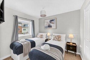 A hotel room with two beds, a window with curtains, a ceiling light, a painting on the wall, and a bedside table with a lamp. - Stylish House • Sleeps 5 • Driveway Parking • Work Desk • Fast WiFi • Self-Check-In • Yoko Property (Northampton)