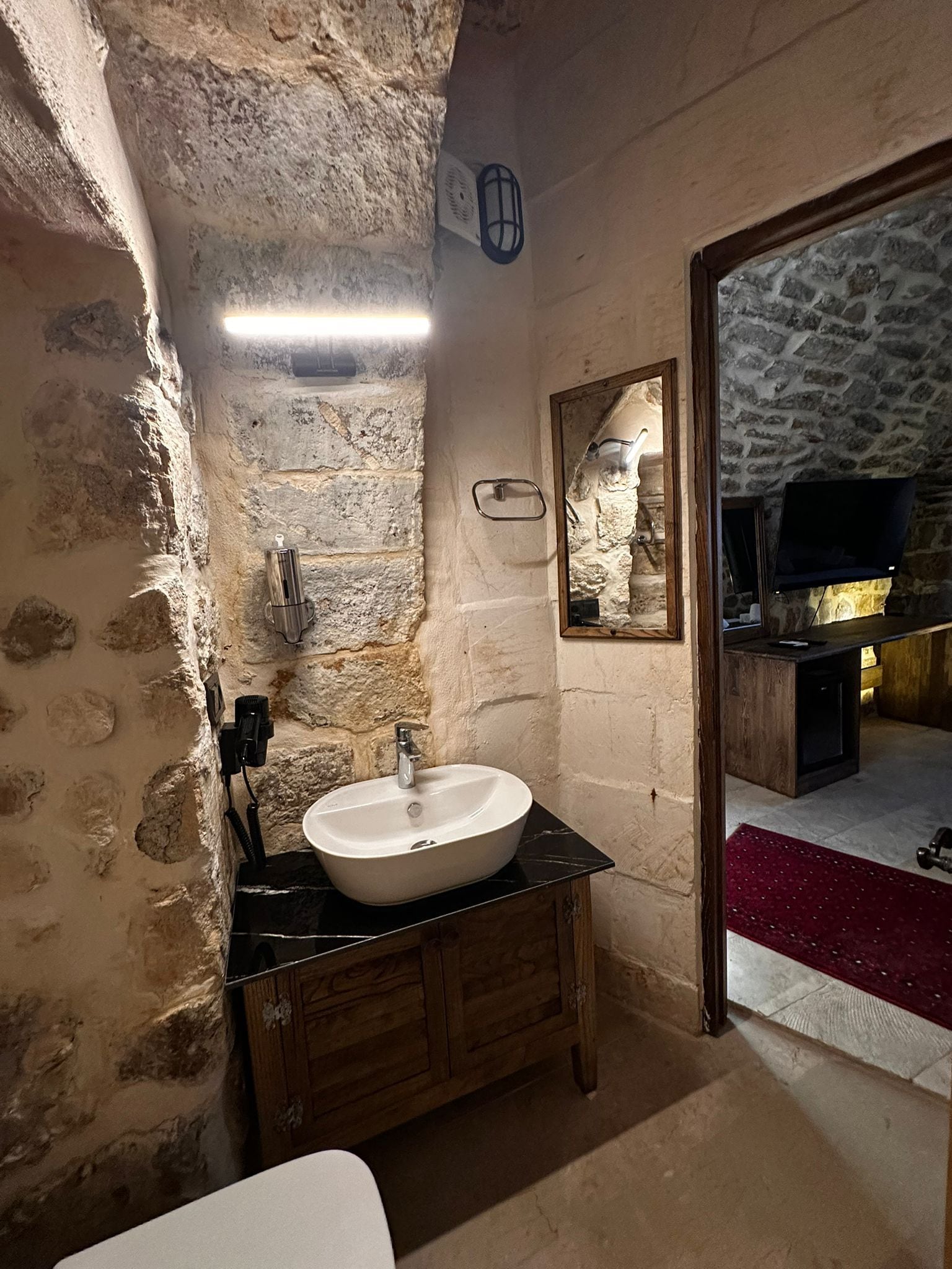 Un baño con pared de piedra, un lavamanos blanco, armarios de madera y un espejo.