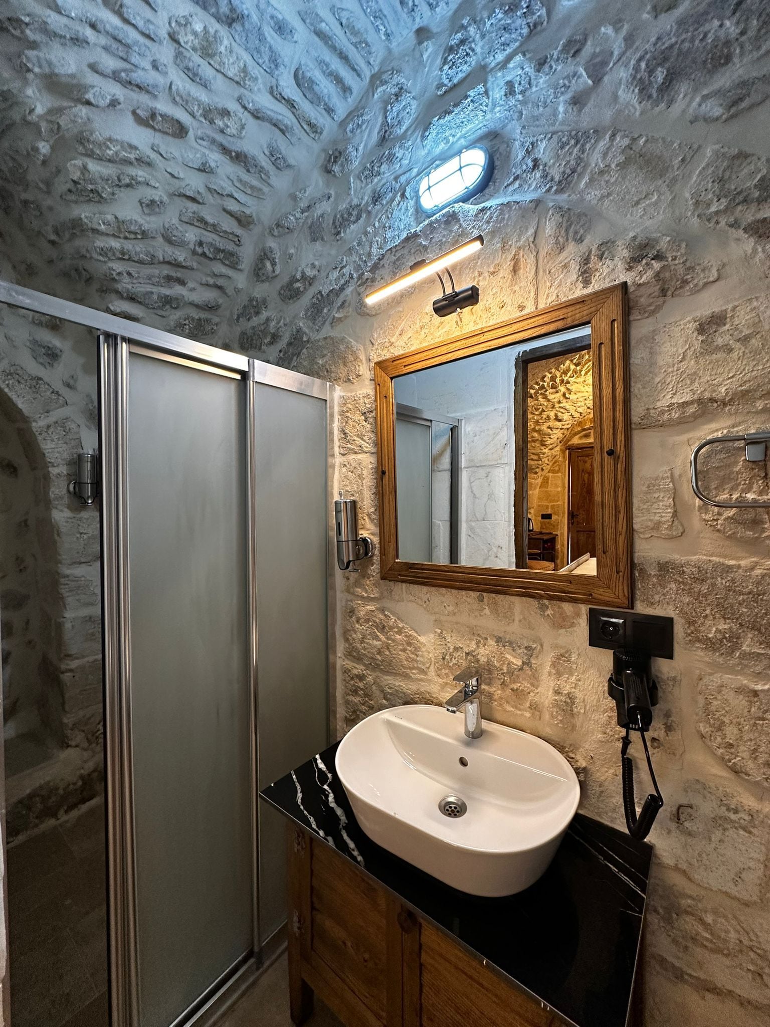 Une salle de bain avec un mur en pierre, un meuble-lavabo en bois, un lavabo blanc et un miroir.