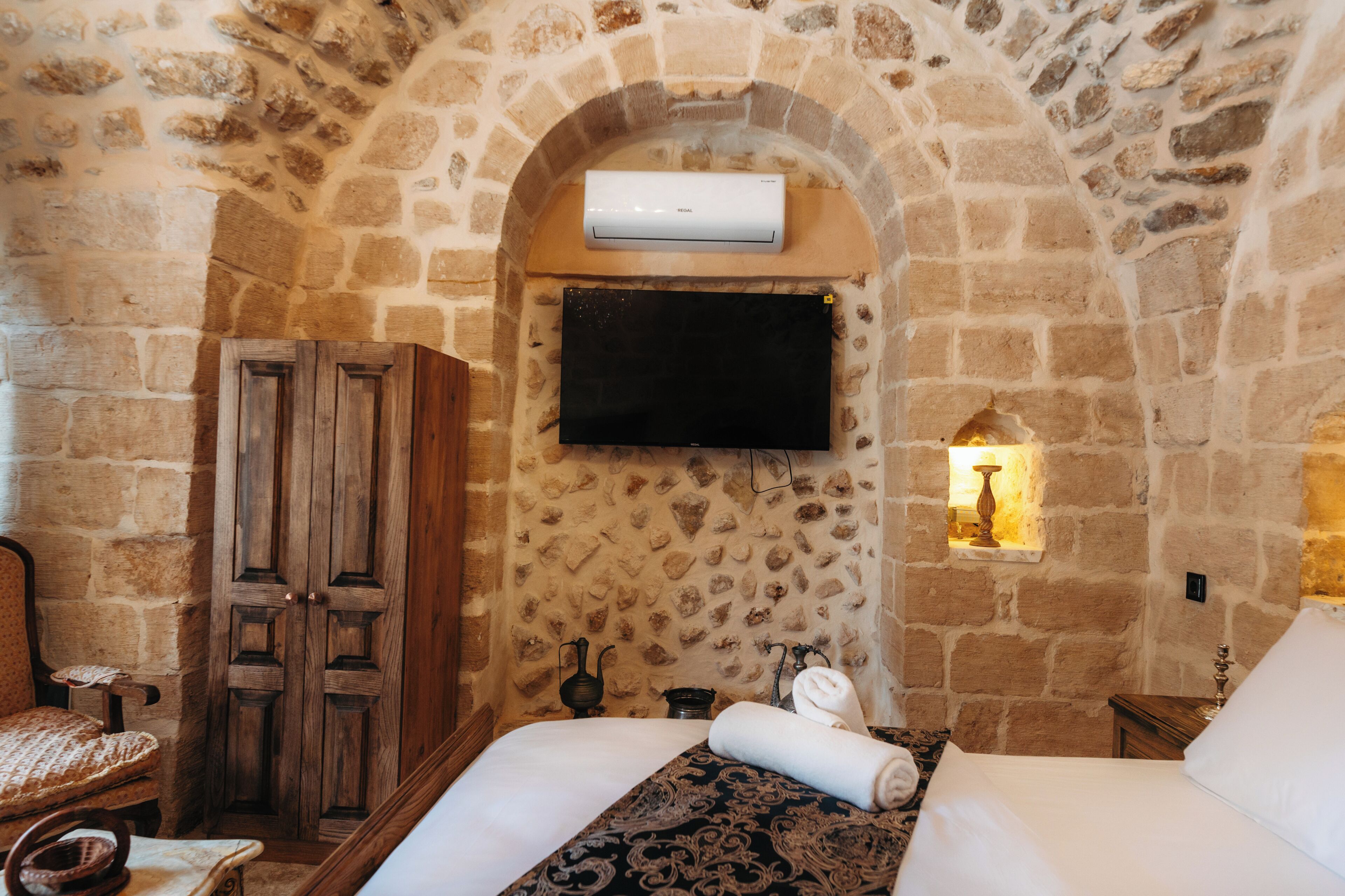 Una habitación con pared de piedra, armario de madera, cama con colcha estampada y televisor de pantalla plana montado en la pared.