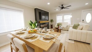Un coin repas avec une table en bois dressée pour le repas, un canapé, un téléviseur et un ventilateur de plafond dans une pièce bien éclairée.