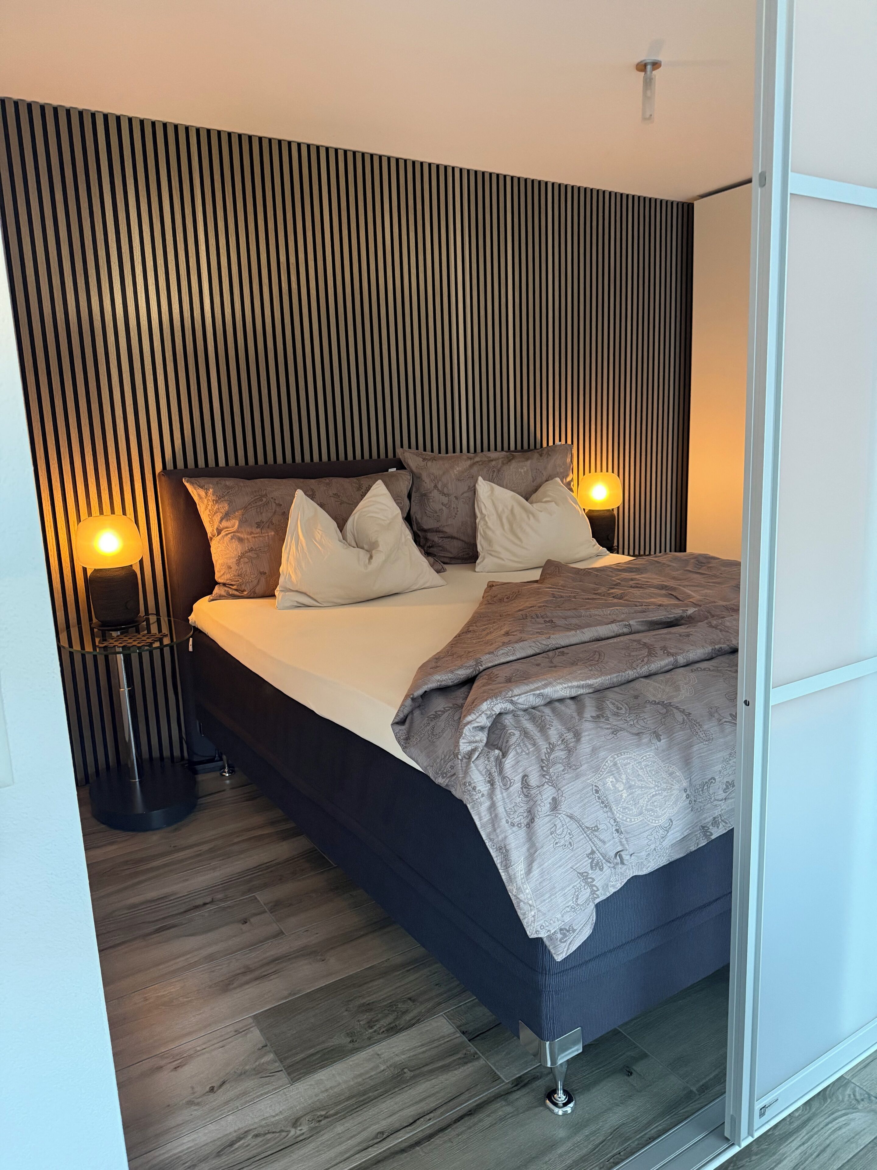 Ein modernes Schlafzimmer mit einem Bett, zwei Nachttischlampen, einem Kopfteil aus Holzleisten und einem Holzboden.